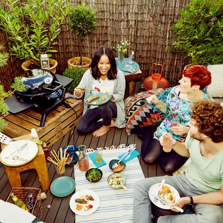 Drei Personen sitzen draußen um einen Tischgrill und genießen eine Mahlzeit Drei Personen sitzen draußen um einen Tischgrill und genießen eine Mahlzeit