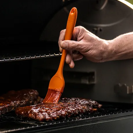 Person bestreicht Steak mit dem Silikonpinsel von Traeger in Orange auf einem Grill. Person bestreicht Steak mit dem Silikonpinsel von Traeger in Orange auf einem Grill.