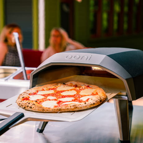 Pizza im Ooni Pizzaofen, im Hintergrund zwei Personen Pizza im Ooni Pizzaofen, im Hintergrund zwei Personen