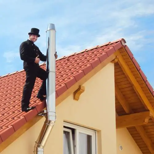 Ramoneur en tenue traditionnelle sur un toit en tuiles à côté d’un conduit en acier inoxydable.