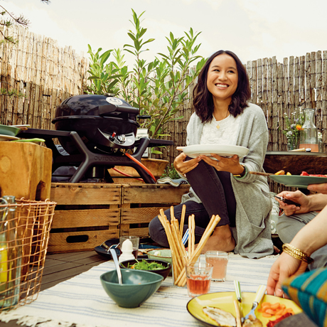 Frau sitzt neben einem Outdoorchef Tischgrill und isst auf einer Terrasse Frau sitzt neben einem Outdoorchef Tischgrill und isst auf einer Terrasse